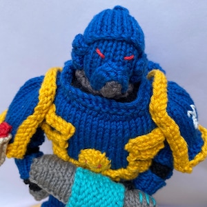 May include: A knitted figurine of a warrior in blue, yellow, and grey. The figure has a blue helmet, yellow shoulder pads, and a grey weapon. Red details are used for the eyes and other accents. The knitted texture is visible throughout the design.