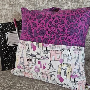 May include: A decorative pillow with a purple floral pattern on top and a white base with a Paris-themed illustration. The pillow has a pocket and is next to a composition notebook and a pen.