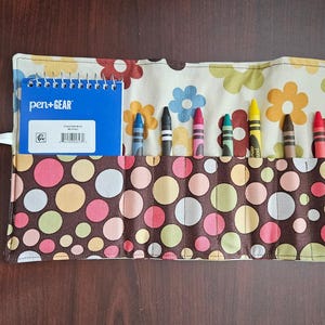 May include: A colorful crayon roll with a floral and polka dot pattern. The roll contains several crayons in various colors and a small blue notebook with the text "pen+GEAR". The roll is tied with a white string.