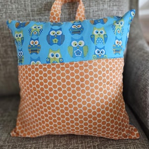 May include: A square pillow with a handle, featuring a blue owl pattern on top and orange polka dots on the bottom. The owls are in shades of blue, green, and yellow. The handle is orange with a polka dot pattern.