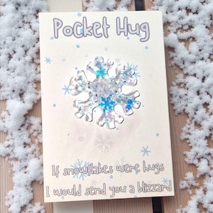 May include: A greeting card with the words "Pocket Hug" and a snowflake design. The snowflake is clear with blue and silver glitter. The card reads, "If snowflakes were hugs I would send you a blizzard."