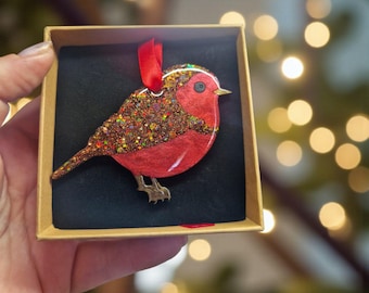 May include: A robin-shaped ornament featuring a red breast and a brown back covered in multi-coloured glitter. The bird has a small gold beak, a black eye, and two gold feet. A red ribbon loop is attached to the top for hanging, presented in a square cardboard box with a black velvet-like insert.