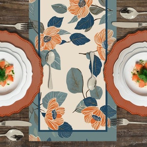 Peut inclure: Un chemin de table beige avec un motif floral bleu et orange. Le chemin de table est décoré de fleurs et de feuilles stylisées. Le chemin de table est placé sur une table en bois avec deux couverts, chacun avec une assiette brune et une assiette blanche.