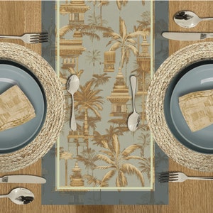May include: A blue and beige table runner with a repeating pattern of palm trees and ornate buildings. The runner is on a wooden table with two place settings, each with a woven placemat and a blue plate.
