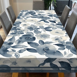 Puede incluir: Un mantel blanco con un patrón floral azul en acuarela. El mantel está sobre una mesa de madera con cuatro sillas.