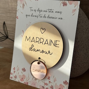 Peut inclure: Une plaque ronde en bois avec les mots "MARRAINE d'amour" et un symbole de cœur. Un petit charme circulaire de couleur or rose avec le nom "Emma" est attaché. La plaque est sur une carte avec des accents floraux et du texte.