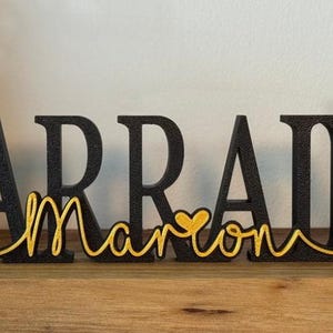 May include: Black wooden letters spelling "MARRAINE" with a gold heart and the word "marron" in gold script.