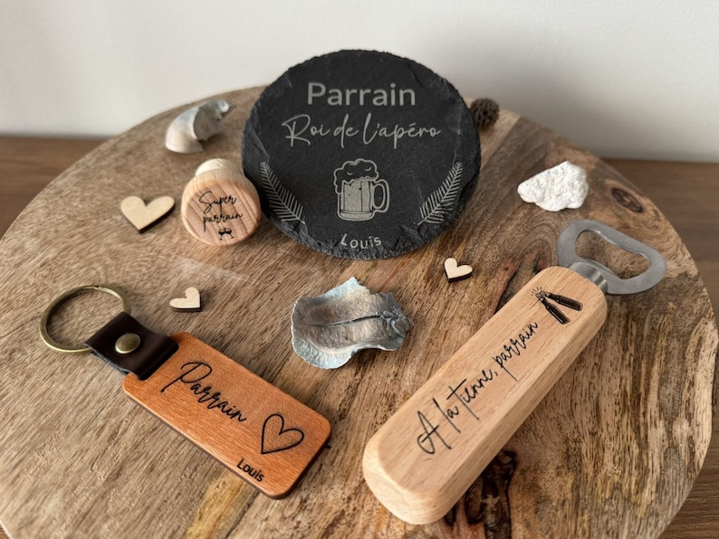 Puede incluir: Un set de regalo de madera para un padrino, que incluye un abridor de botellas con el texto "A ta time, parrain", un llavero con el texto "Parrain" y un coraz&oacute;n, y un posavasos de pizarra con el texto "Parrain Roi de l'ap&eacute;ro" y una jarra de cerveza.