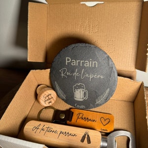 Puede incluir: Un set de regalo para un padrino, que incluye un posavasos de pizarra con el texto "Parrain Roi de l'ap&eacute;ro" y un gr&aacute;fico de jarra de cerveza, un abridor de botellas de madera con el texto "A la tienne, parrain" y un gr&aacute;fico de dos botellas de cerveza, un tap&oacute;n de botella de madera con el texto "Super parrain", y un llavero de cuero con el texto "Parrain" y un coraz&oacute;n.