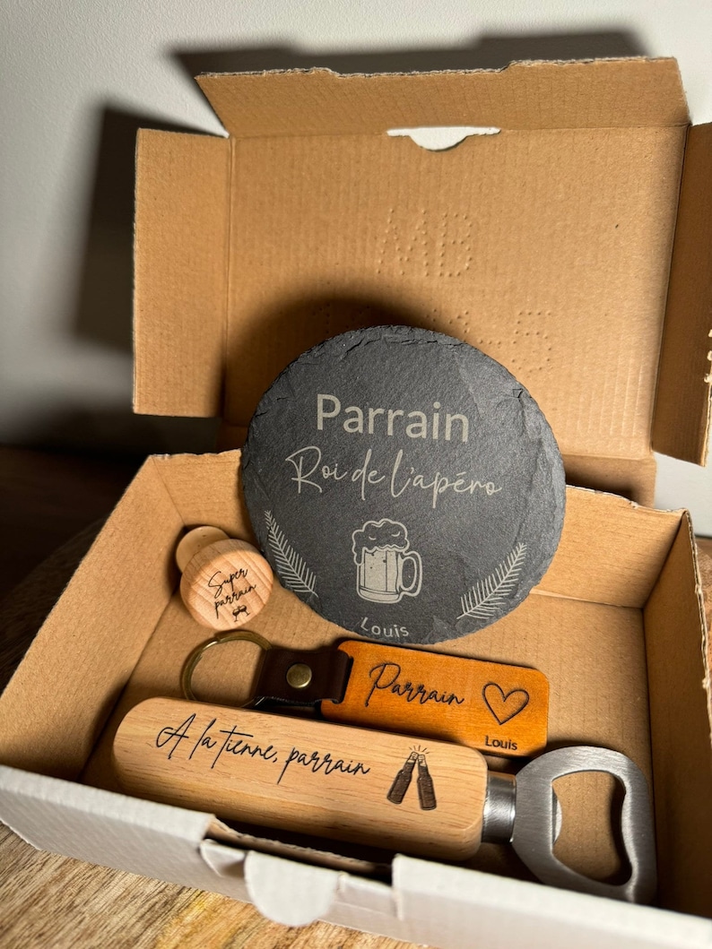 Puede incluir: Un set de regalo para un padrino, que incluye un posavasos de pizarra con el texto "Parrain Roi de l'ap&eacute;ro" y un gr&aacute;fico de jarra de cerveza, un abridor de botellas de madera con el texto "A la tienne, parrain" y un gr&aacute;fico de dos botellas de cerveza, un tap&oacute;n de botella de madera con el texto "Super parrain", y un llavero de cuero con el texto "Parrain" y un coraz&oacute;n.