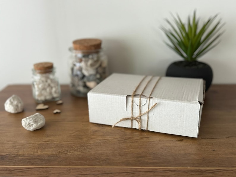 Puede incluir: Una caja de cart&oacute;n blanca atada con cuerda, sentada sobre una mesa de madera. Dos frascos de vidrio con tapas de corcho llenos de piedras blancas est&aacute;n en el fondo.