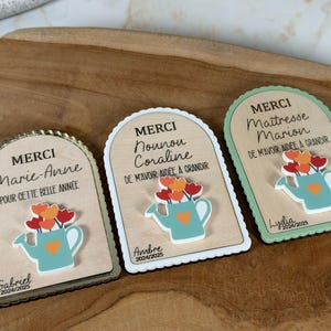 Peut inclure: Trois plaques en bois avec un motif d'arrosoir et le mot "MERCI" en haut. Chaque plaque porte un nom et un message différents, avec les années 2024/2025 en bas. Les arrosoirs sont bleu clair avec des fleurs orange et rouges.