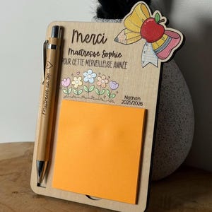 Peut inclure: Un support de bloc-notes en bois clair avec un stylo en bambou et un bloc de notes adhésives orange vif. Le bois est gravé avec le texte "Merci Maitresse Sophie POUR CETTE MERVEILLEUSE ANNÉE", "Nathan 2025/2026" et "Maitresse au top".