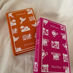 Può includere: Due libri rilegati con copertine arancioni e rosa. Il libro arancione ha il titolo "Wild Eyes" e il libro rosa ha il titolo "Wild Love". Entrambi i libri hanno illustrazioni e testo bianchi sulle copertine.