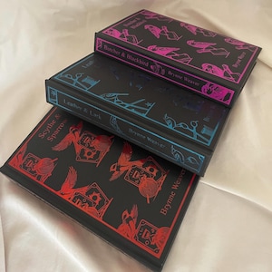 Può includere: Tre libri rilegati impilati. Il libro in alto è nero con illustrazioni rosa e il titolo "Butcher & Blackbird". Il libro centrale è nero con illustrazioni blu e il titolo "Leather & Lark". Il libro in basso è nero con illustrazioni rosse e il titolo "Scythe & Sparrow".