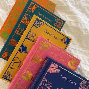 Può includere: Una pila di sei libri rilegati in tela colorati con lettere dorate e illustrazioni. I titoli dei libri sono "People We Meet", "Book Lovers", "Beach Read", "Happy Place", "Funny Story" e "Cherry Hill Winery".