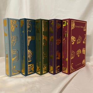 Può includere: Una collezione di sei libri con copertina rigida e dettagli in lamina d'oro. I libri hanno dorsi di diversi colori, tra cui verde acqua, verde e bordeaux. I titoli e il nome dell'autore sono stampati.