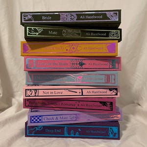 Può includere: Una pila di libri colorati di Ali Hazelwood. Le copertine mostrano titoli come "Bride", "Mate" e "The Love Hypothesis". Ogni libro ha uno schema di colori unico, con testo ed elementi decorativi.