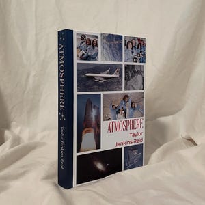 Può includere: Un libro con copertina rigida intitolato "Atmosphere" di Taylor Jenkins Reid. La copertina presenta un collage di immagini, tra cui foto di astronauti, un lancio di razzi e una galassia. Il dorso è blu scuro con il titolo e il nome dell'autore.