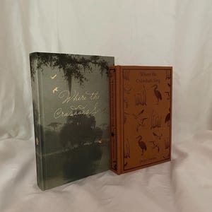 Può includere: Sono esposti due libri con copertina rigida, "Where the Crawdads Sing". Uno ha una copertina verde con un albero e scritte dorate. L'altro ha una copertina marrone con illustrazioni di uccelli e il titolo.