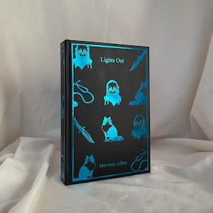 Può includere: Un libro rilegato nero con un design in lamina blu. Il titolo "Lights Out" è stampato sulla copertina, insieme al nome dell'autore "Navessa Allen".