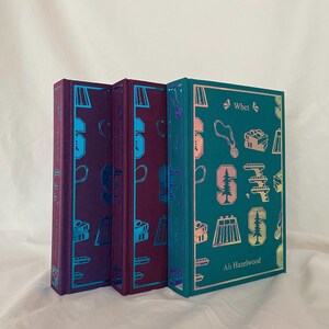 Può includere: Tre libri rilegati con copertine verde acqua, viola e blu. Il libro a destra ha il titolo "Whet" e l'autore "Ali Hazelwood" stampato sulla copertina.