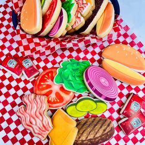 May include: A collection of decorated sugar cookies shaped like hamburger ingredients, including buns, lettuce, tomato, onion, pickles, bacon, cheese, and a burger patty. The cookies are arranged on a red and white checkered surface, with ketchup packets.