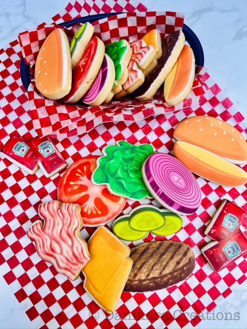 May include: A collection of decorated sugar cookies shaped like hamburger ingredients, including buns, lettuce, tomato, onion, pickles, bacon, cheese, and a burger patty. The cookies are arranged on a red and white checkered surface, with ketchup packets.