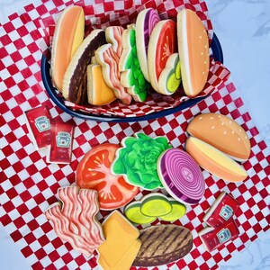 May include: A collection of decorated sugar cookies shaped like a hamburger with all the fixings, including a bun, patty, cheese, lettuce, tomato, onion, bacon, and pickles. Two ketchup packets are also included. The cookies are arranged on a red and white checkered paper.