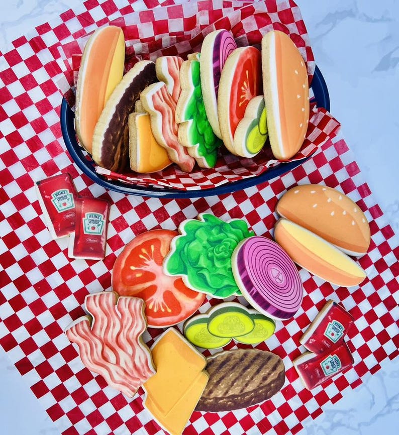 May include: A collection of decorated sugar cookies shaped like a hamburger with all the fixings, including a bun, patty, cheese, lettuce, tomato, onion, bacon, and pickles. Two ketchup packets are also included. The cookies are arranged on a red and white checkered paper.