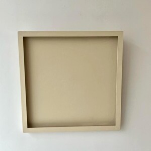 May include: A square, beige-colored tray with a raised border. The tray is a solid color and appears to be made of wood or a similar material. The tray is empty and sits against a white wall.