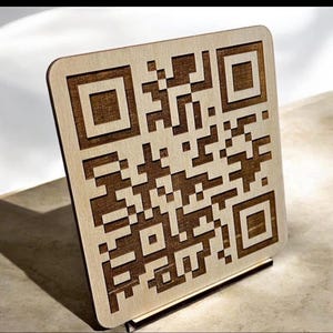 May include: A square wooden sign featuring a laser-engraved QR code pattern. The design consists of a light-colored wood surface with dark brown, recessed square and rectangular pixelated shapes forming a functional scannable code pattern.