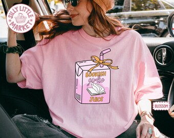 May include: Pale pink t-shirt with a juice box design. The juice box graphic reads "Bookish Girlie Juice" and includes an open book illustration, a straw, and a yellow bow. The shirt is a light pink colour.