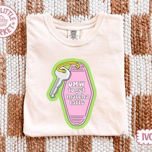 May include: Ivory-colored t-shirt with a graphic of a key and a motel key tag that reads "OMW to get matcha latte." The key tag is pink with a green border. The shirt is folded on a woven brown and white surface.