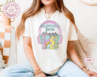 Può includere: T-shirt color avorio con la scritta "Audiobook Girlie" sopra una grafica di cuffie e libri. I libri presentano disegni arcobaleno, floreali e a quadri. La maglietta ha maniche corte e girocollo.