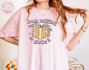 May include: A pale pink t-shirt featuring the words "Book sniffing girlie" above a cartoon book illustration. The book is yellow and pink checkered with pink bows. The shirt has the "Silly Little Market" logo.