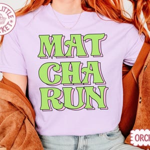 May include: A light purple t-shirt with the words "MAT CHA RUN" in green letters with a pink outline. The shirt has a crew neck and short sleeves. The text "ORCHID" is in a pink oval on the bottom right.
