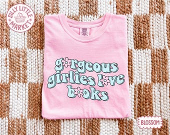 May include: A pale pink t-shirt featuring the phrase "gorgeous girlies love books" in a retro font with light blue detailing. The shirt is made of a soft fabric and is displayed flat on a patterned surface. The shirt is labelled "Blossom".