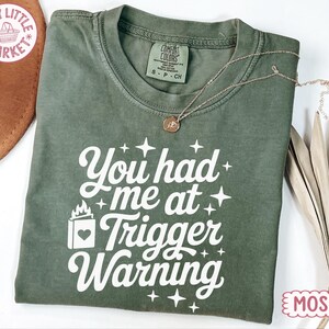 May include: A moss green short-sleeve t-shirt featuring white screen-printed text that reads You had me at Trigger Warning, accompanied by a small book icon with a heart and flame, surrounded by several four-pointed star graphics.