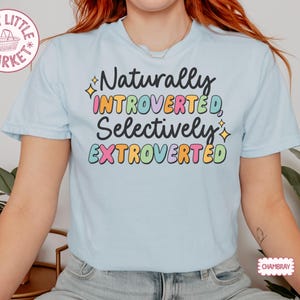 May include: Light blue t-shirt with the text "Naturally Introverted, Selectively Extroverted" in colorful bubble letters. The shirt has short sleeves and a crew neck. The design includes star accents.