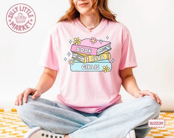 May include: A pale pink t-shirt with a graphic of three books stacked on top of each other. The books have the words "BOOK CLUB GIRLIE" on them. The design includes yellow flowers and stars. The person is sitting cross-legged.