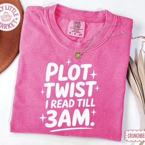 May include: A pink short-sleeved t-shirt with white text that reads PLOT TWIST I READ TILL 3AM. The shirt features a crew neckline, a Comfort Colors brand label, and small white star graphics surrounding the text on the front of the garment.