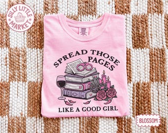 Può includere: T-shirt rosa chiaro con la scritta "SPREAD THOSE PAGES LIKE A GOOD GIRL" e una grafica di libri, un bicchiere di vino e altri elementi. La maglietta è del marchio Comfort Colors.