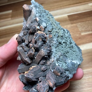 Può includere: Un gruppo di cristalli marrone scuro e grigi con una superficie ruvida e strutturata. I cristalli sono raggruppati, con alcune aree che mostrano un colore più chiaro, quasi traslucido. L'aspetto generale è naturale e geologico.