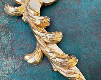 May include: A gold-colored, ornate decorative architectural element with a leaf-like design. The piece has a curved shape with detailed, layered textures and a distressed finish, showing hints of silver. The object is likely made of wood or a similar material.