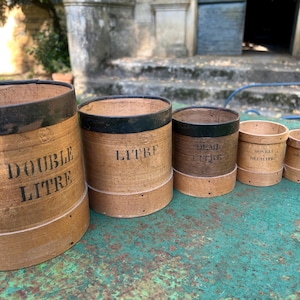 Op de afbeelding: Een set van zes antieke houten maatbekers. De bekers zijn cilindrisch met zwarte metalen banden en voorzien van zwarte tekst die het volume aangeeft, waaronder "DOUBLE LITRE" en "LITRE". Ze zijn gerangschikt op een groene ondergrond.