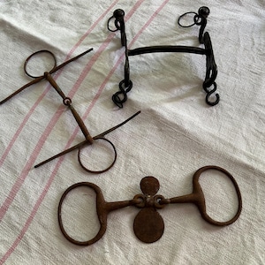 Op de afbeelding: Drie antieke paardengebitten in verschillende ontwerpen, gemaakt van donker metaal. Eén bit heeft een rechte staaf met ringen, een ander heeft een gebogen staaf met decoratieve krullen en de derde heeft een centrale plaat. De bitten liggen op een wit en rood gestreepte doek.