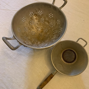 Könnte beinhalten: Zwei vintage Aluminium-Siebe, eines mit zwei Griffen und das andere mit einem Holzgriff. Beide Siebe haben ein perforiertes Design.