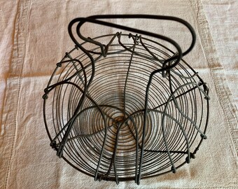 Puede incluir: Una cesta de alambre vintage con asa. La cesta está hecha de alambre negro y tiene forma circular. Está diseñada para contener fruta u otros artículos.