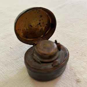 Pode incluir: Um tinteiro antigo, de cor bronze, com tampa articulada. O tinteiro tem uma base redonda e um pequeno tinteiro elevado. A tampa está aberta, revelando o interior. O tinteiro é provavelmente feito de latão ou metal semelhante.